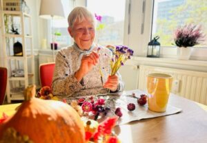 Ältere Frau sitzt an einem Tisch und arrangiert Blumen, mit einem Kürbis im Vordergrund.