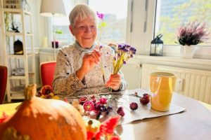 Ältere Frau sitzt an einem Tisch und arrangiert Blumen, mit einem Kürbis im Vordergrund.