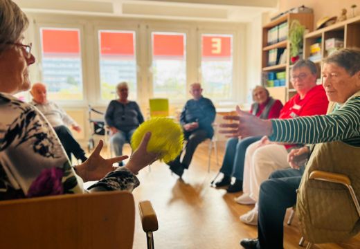 Eine Gruppe von Senioren sitzt im Kreis in einem Raum, während eine Person einen gelben Ball weiterreicht.