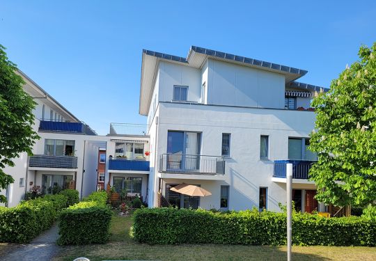 Moderne Wohnanlage mit mehreren Etagen, umgeben von grünen Büschen und Bäumen bei sonnigem Wetter.