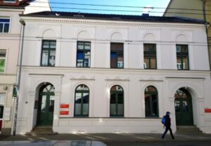 Ein zweistöckiges Gebäude mit weißer Fassade und grünen Fensterrahmen. Vor dem Gebäude geht eine Person vorbei.