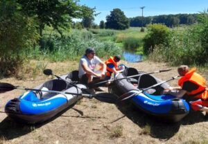 Drei Personen sitzen in blauen aufblasbaren Kajaks am Ufer eines kleinen Gewässers.