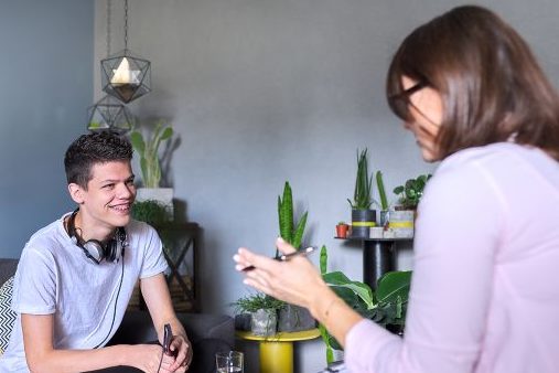Jugendlicher sitzt auf einem Sofa und unterhält sich mit einer Frau, die ihm gegenüber sitzt.