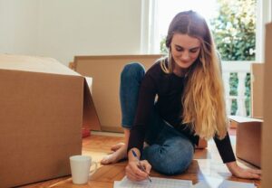 Junge Frau sitzt auf dem Boden zwischen Umzugskartons und schreibt auf ein Papier.