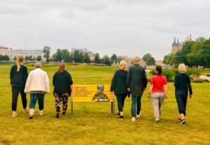 Eine Gruppe von sieben Menschen geht über eine Wiese in einem Park mit einem gelben Schild im Vordergrund.
