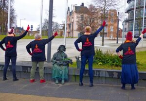 Vier Personen stehen mit dem Rücken zur Kamera neben einer Statue einer sitzenden Frau. Sie tragen schwarze Jacken mit roten Symbolen und heben den rechten Arm in die Luft.