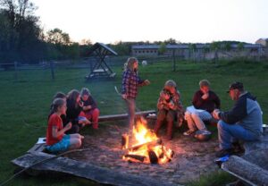 Kinder und Erwachsene sitzen um ein Lagerfeuer auf einer Wiese