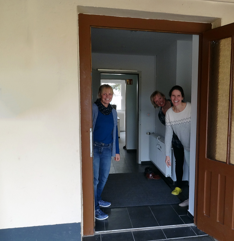 Drei Frauen stehen lächelnd in einem Eingangsbereich und schauen aus der Tür heraus.