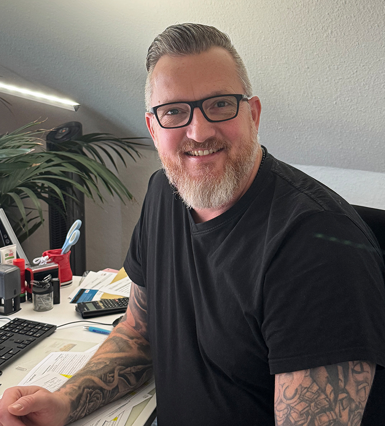 Ein lächelnder Mann mit Brille und Bart sitzt an einem Schreibtisch in einem Büro.