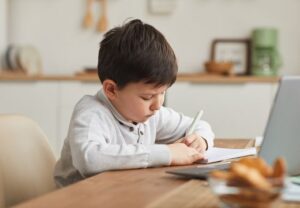 Ein kleiner Junge sitzt an einem Tisch und schreibt konzentriert in ein Heft.