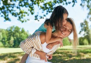 Eine Frau trägt ein lächelndes junges Mädchen auf ihrem Rücken in einem sonnigen Park.