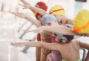 Kinder mit Schwimmkappen stehen am Beckenrand und strecken die Arme aus