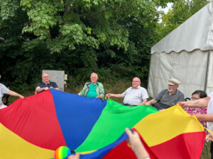 Eine Gruppe von älteren Menschen sitzt im Freien und hält ein buntes Fallschirmtuch.