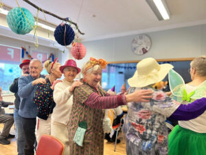 Ältere Menschen nehmen an einer Feier teil und tragen festliche Kleidung und Hüte, einige mit dekorativen Accessoires.
