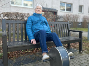 Ältere Person sitzt auf einer Parkbank und nutzt ein Tretgerät.