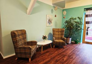 Ein gemütlicher Raum mit zwei karierten Sesseln, einem kleinen Tisch mit Blumenstrauß, hängender Dekoration, einer Pflanze und einem Gemälde an der Wand.