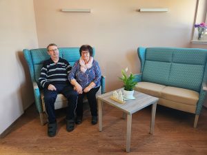 Zwei ältere Personen sitzen in einem gemütlich eingerichteten Raum auf einem blauen Sofa.