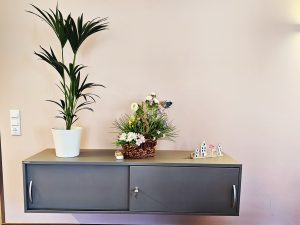 Ein Sideboard mit einer großen Topfpflanze, einem Blumenkorb und kleinen Deko-Häusern.