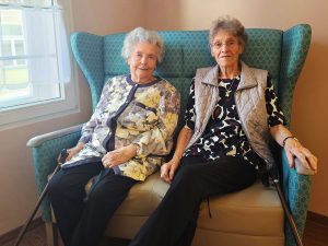 Zwei ältere Frauen sitzen auf einem blauen Sessel in einem Raum mit Fenster.