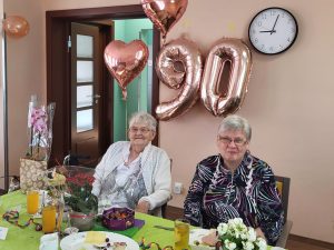 Zwei ältere Frauen sitzen an einem Tisch, der für eine Feier dekoriert ist, mit Ballons in Form der Zahl 90 im Hintergrund.