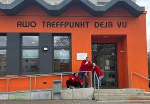 Drei Personen in roten Oberteilen vor dem Eingang des AWO Treffpunkts „Deja Vu“ in einem orangefarbenen Gebäude mit Rampe und Treppe.