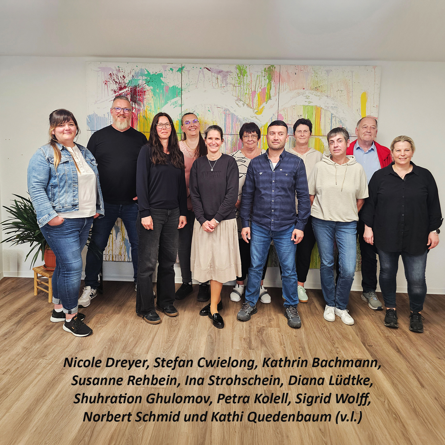 Eine Gruppe Menschen in einem Raum vor einem farbenfrohen Kunstwerk an der Wand.