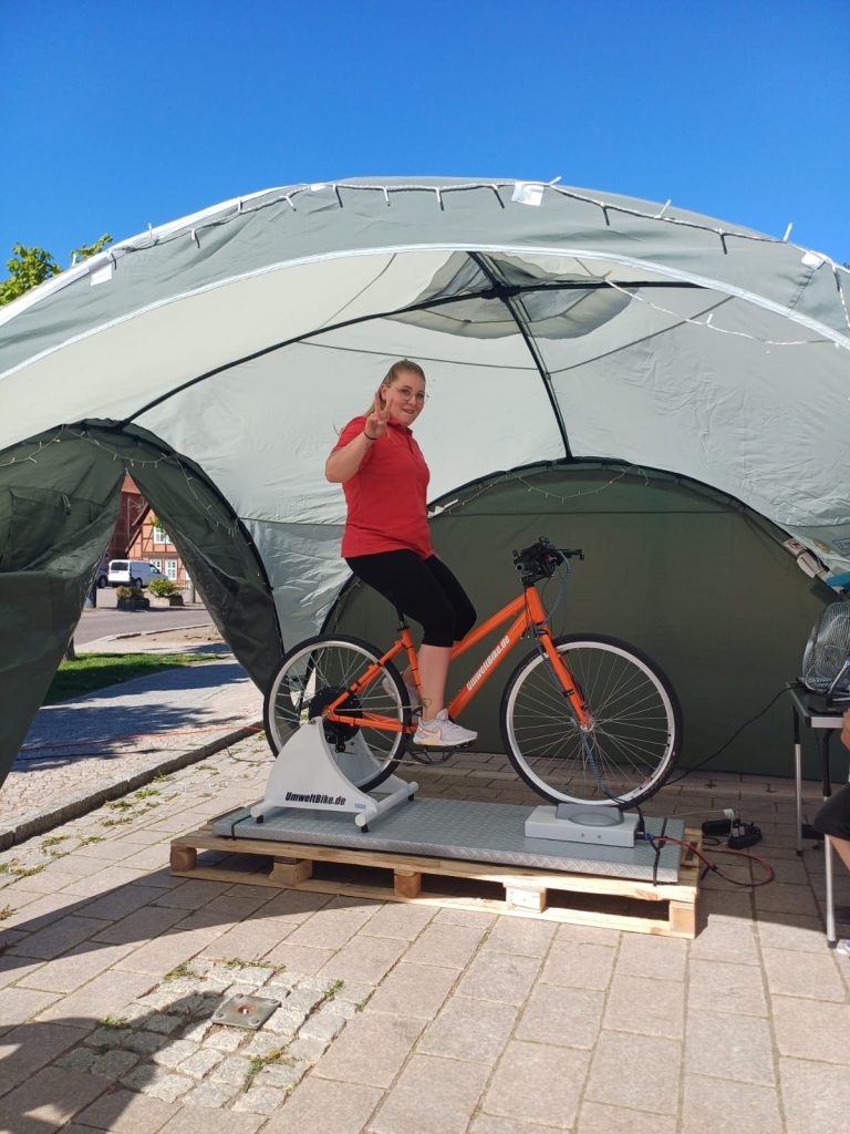 Person auf einem Standfahrrad, l&auml;chelnd und das Peace-Zeichen zeigend, unter einem gr&uuml;nen Zelt.