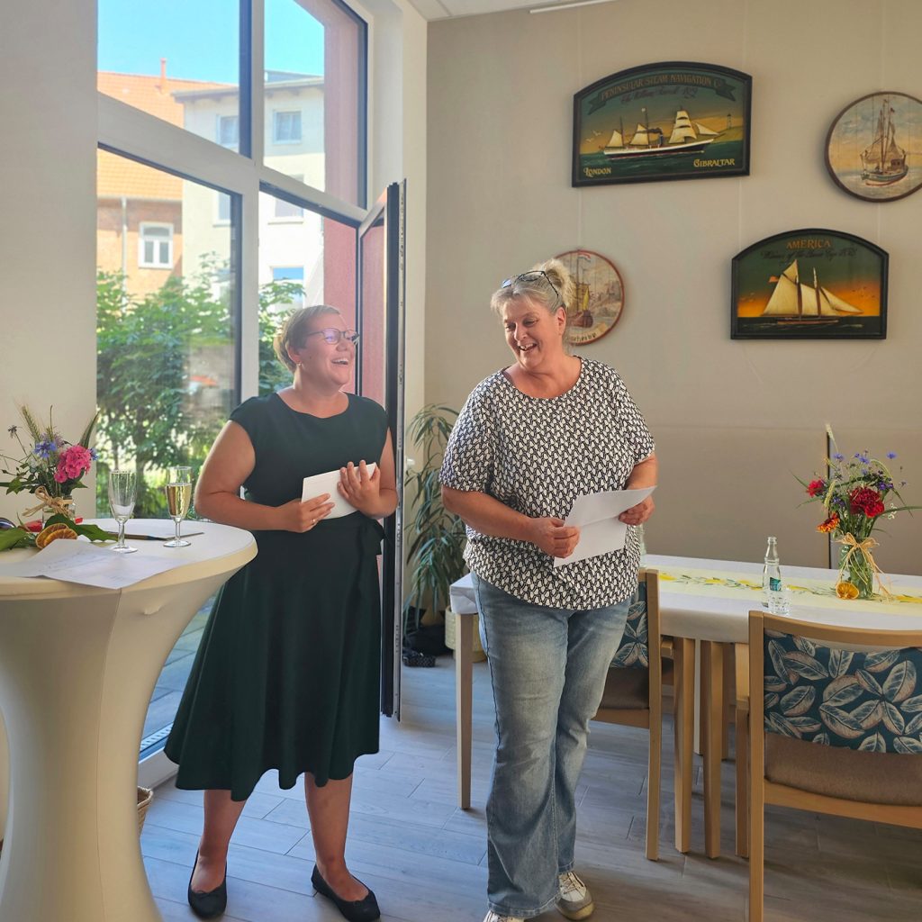Zwei Frauen stehen l&auml;chelnd in einem Raum neben einem Stehtisch mit Blumen und Getr&auml;nken.