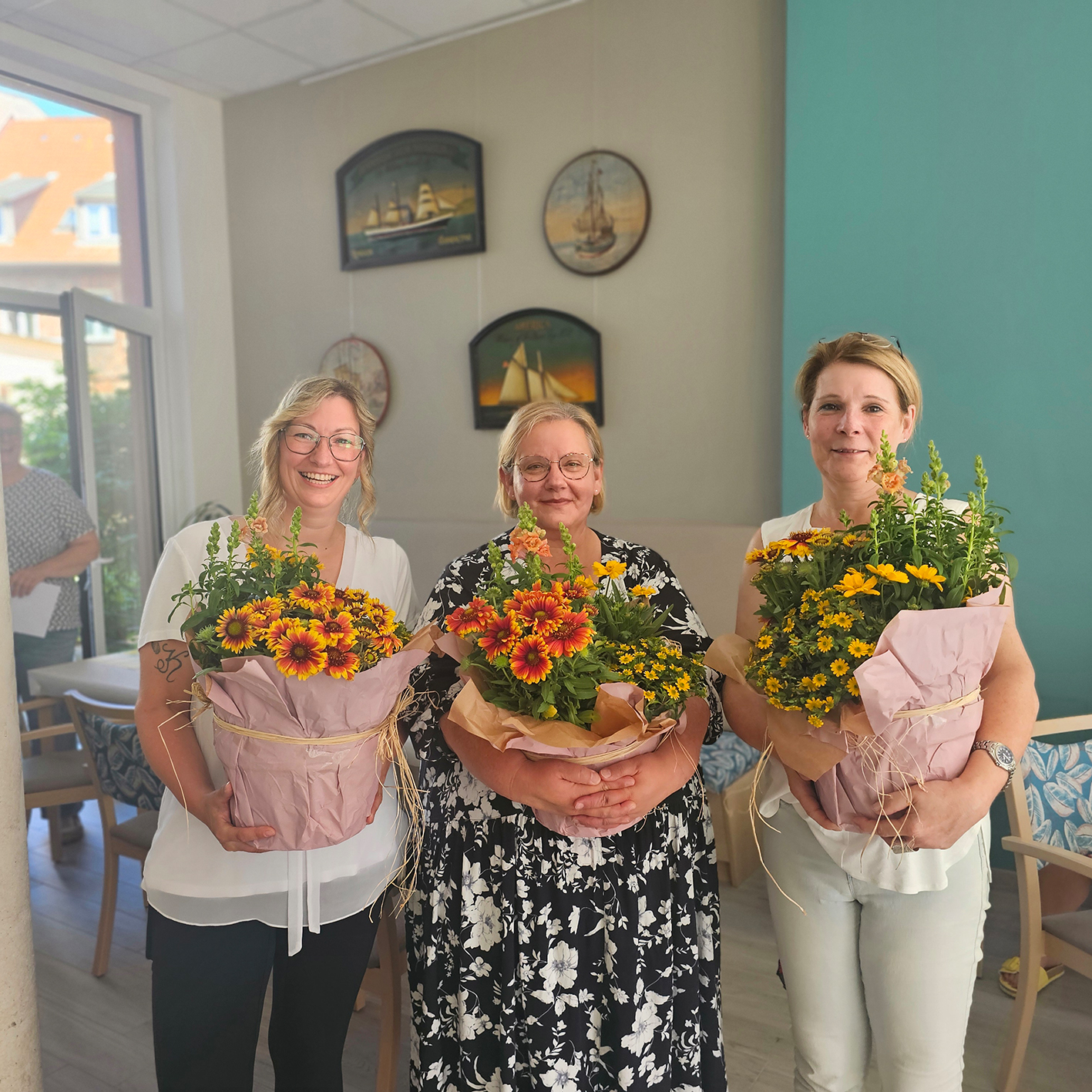 Drei Frauen halten bunte Blumengestecke in einem Raum mit maritimer Dekoration.