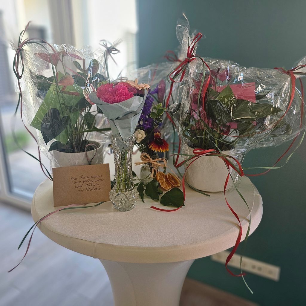 Verschiedene Blumenarrangements stehen auf einem wei&szlig; gedeckten Tisch, darunter ein Blumenstrau&szlig; in einer Vase sowie Blument&ouml;pfe in Klarsichtfolie verpackt, verziert mit Schleifen.