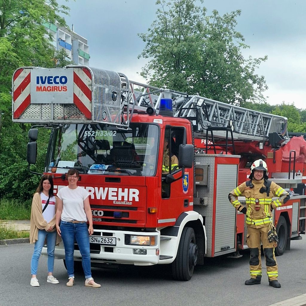 Ein rotes Feuerwehrfahrzeug mit zwei Frauen und einem Feuerwehrmann davor.