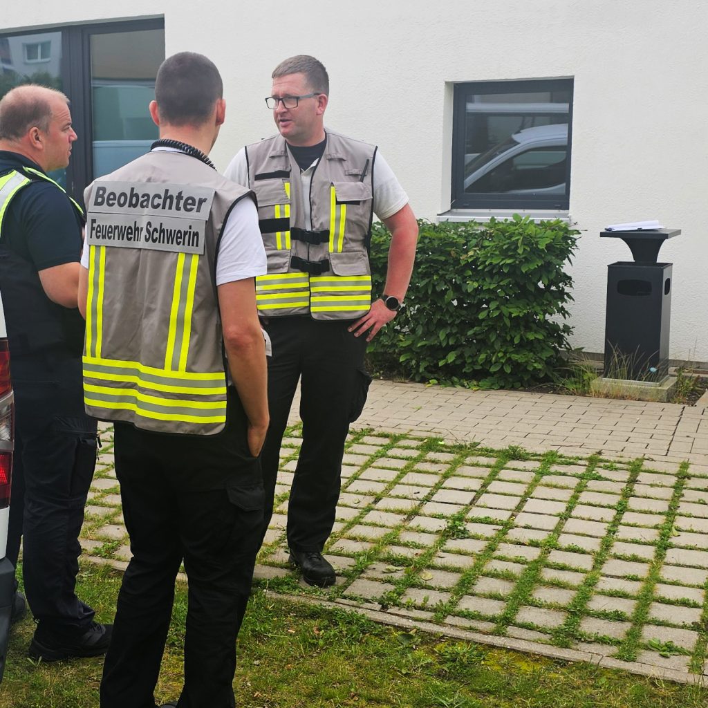 Drei M&auml;nner in Feuerwehr-Westen neben einem Geb&auml;ude