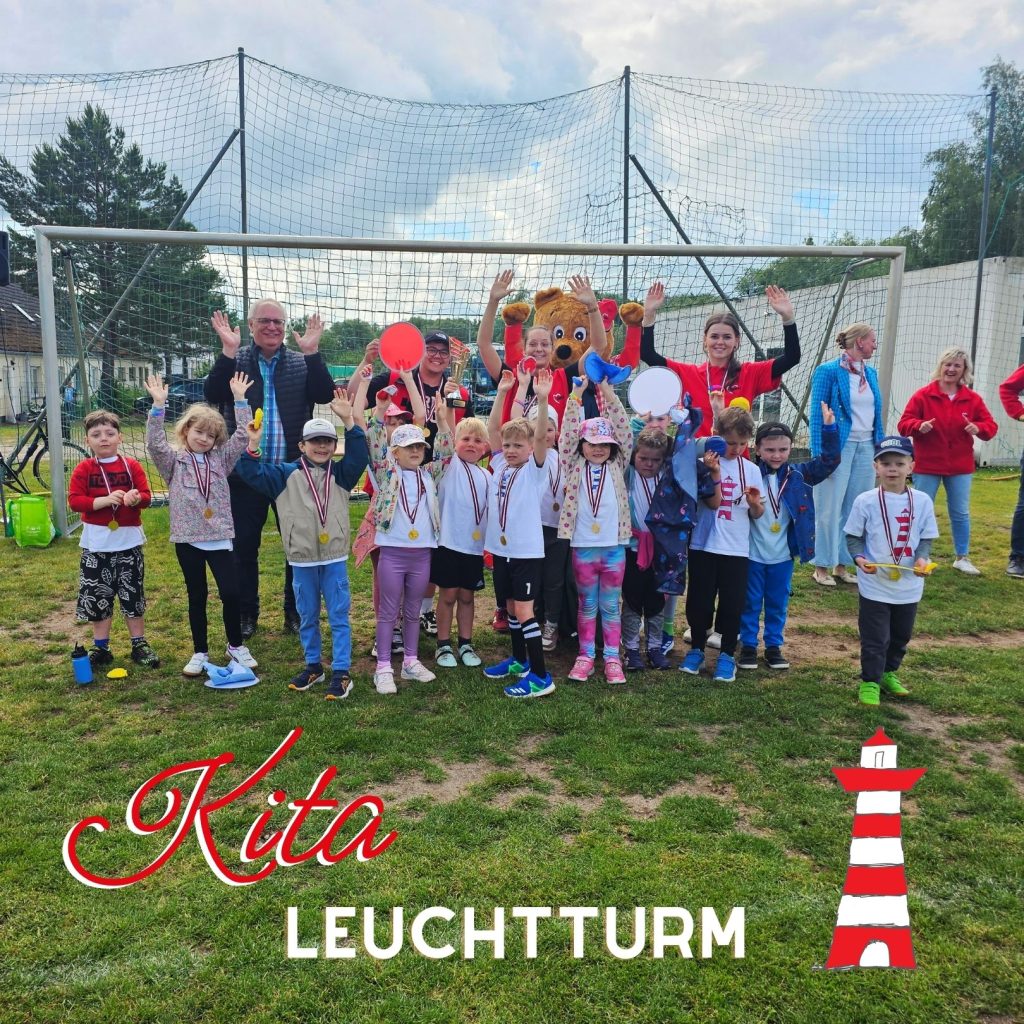 Eine Gruppe von Kindern und Erwachsenen steht unter einem Fu&szlig;balltor auf einem Rasenplatz und feiert. Einige der Personen halten Luftballons, w&auml;hrend die Kinder Medaillen um den Hals tragen. Der Name 'Kita Leuchtturm' ist in roter und wei&szlig;er Schrift in der linken unteren Ecke zu sehen, begleitet von einer Grafik eines Leuchtturms.