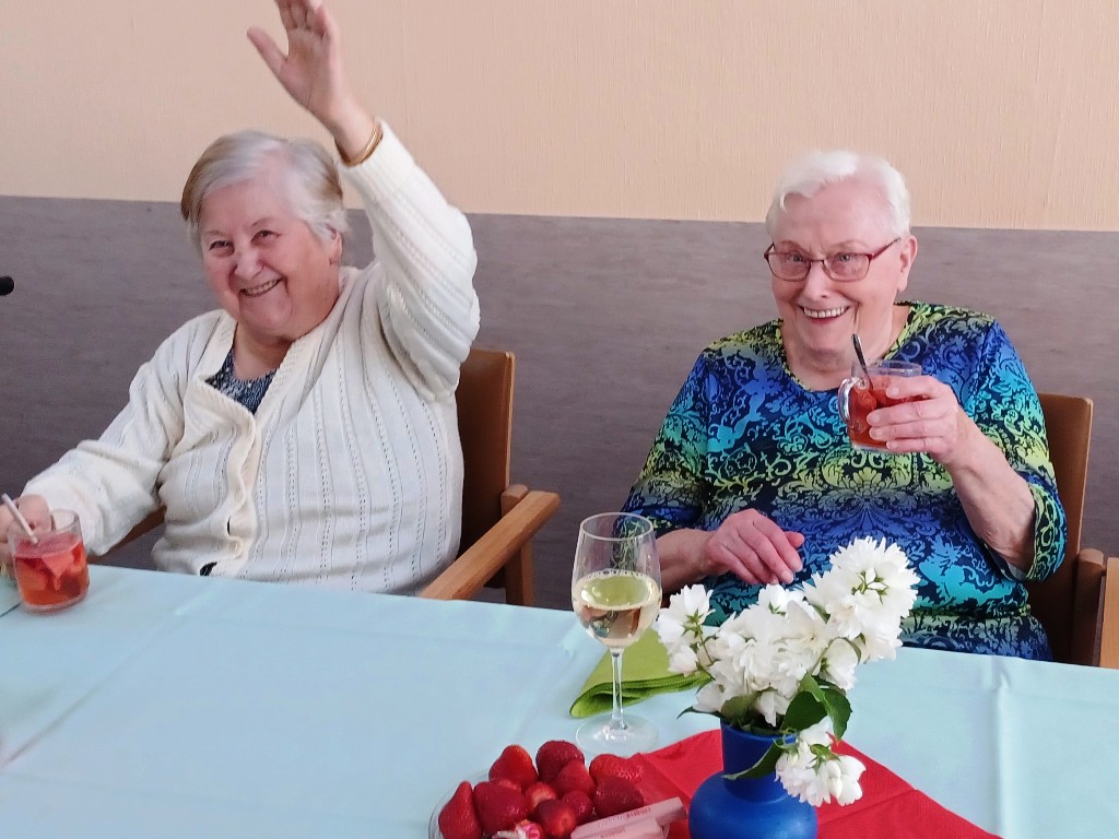 Zwei lächelnde ältere Frauen sitzen an einem Tisch mit Getränken und Blumen.