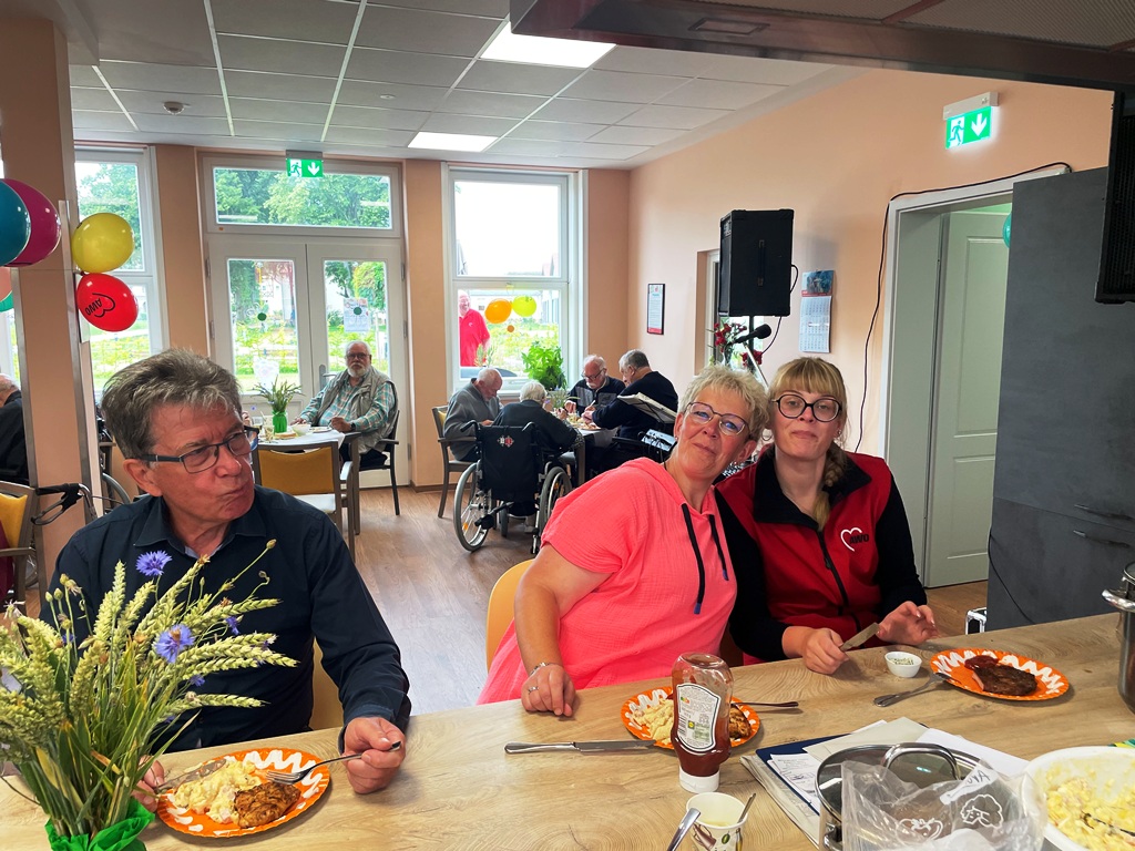 Drei Personen sitzen an einem Tisch und essen, im Hintergrund sind weitere Menschen zu sehen.
