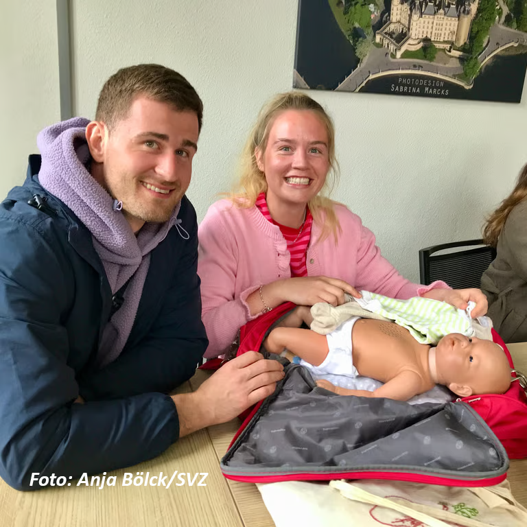 Ein Mann und eine Frau sitzen an einem Tisch und lächeln in die Kamera. Vor ihnen liegt eine Babypuppe in einer Tasche.