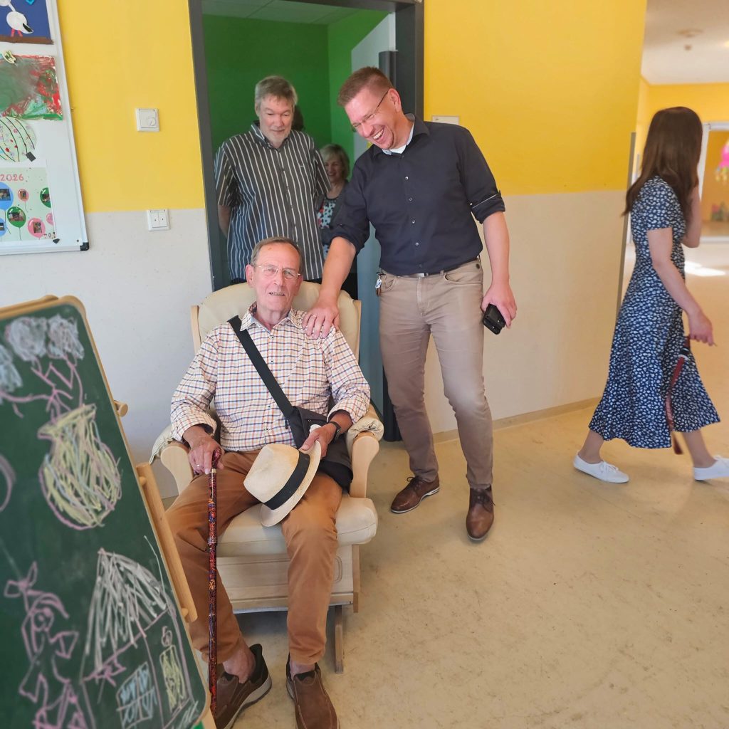 Eine Person sitzt auf einem Stuhl, umgeben von anderen Personen in einem Raum mit gelben Wänden.