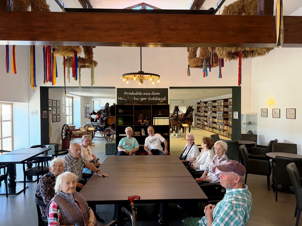 Gruppe älterer Menschen in einem Gemeinschaftsraum mit traditionellen Dekorationen und einem Bücherregal im Hintergrund.