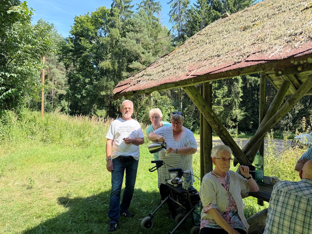 Gruppe von Menschen unter einem hölzernen Unterstand im Grünen.