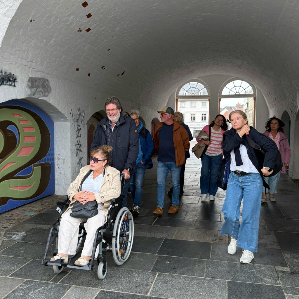 Gruppe von Menschen, darunter eine Person im Rollstuhl, in einem überdachten Durchgang.
