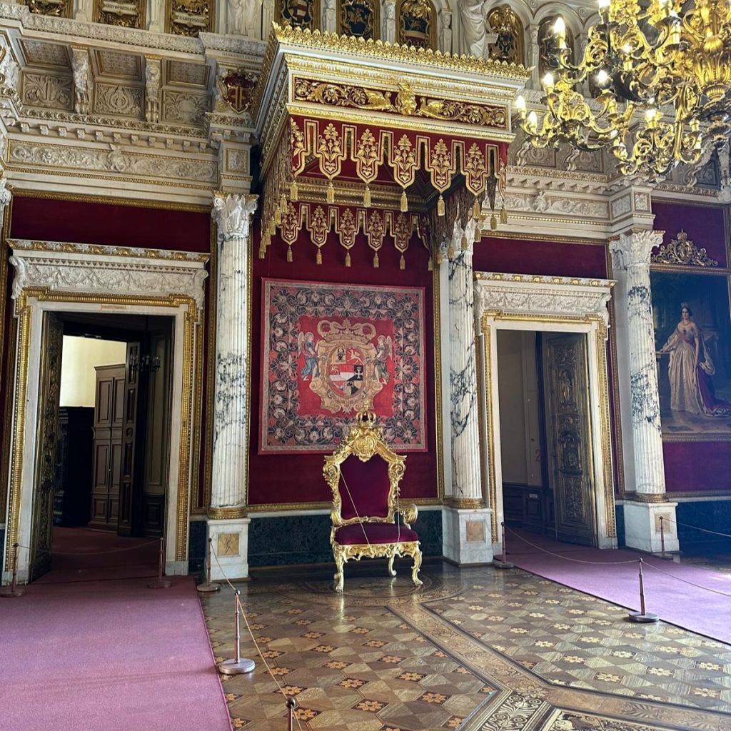 Prunkvoller Thronsaal mit einem reich verzierten Thron, roten Teppichen und goldenen Dekorationen.