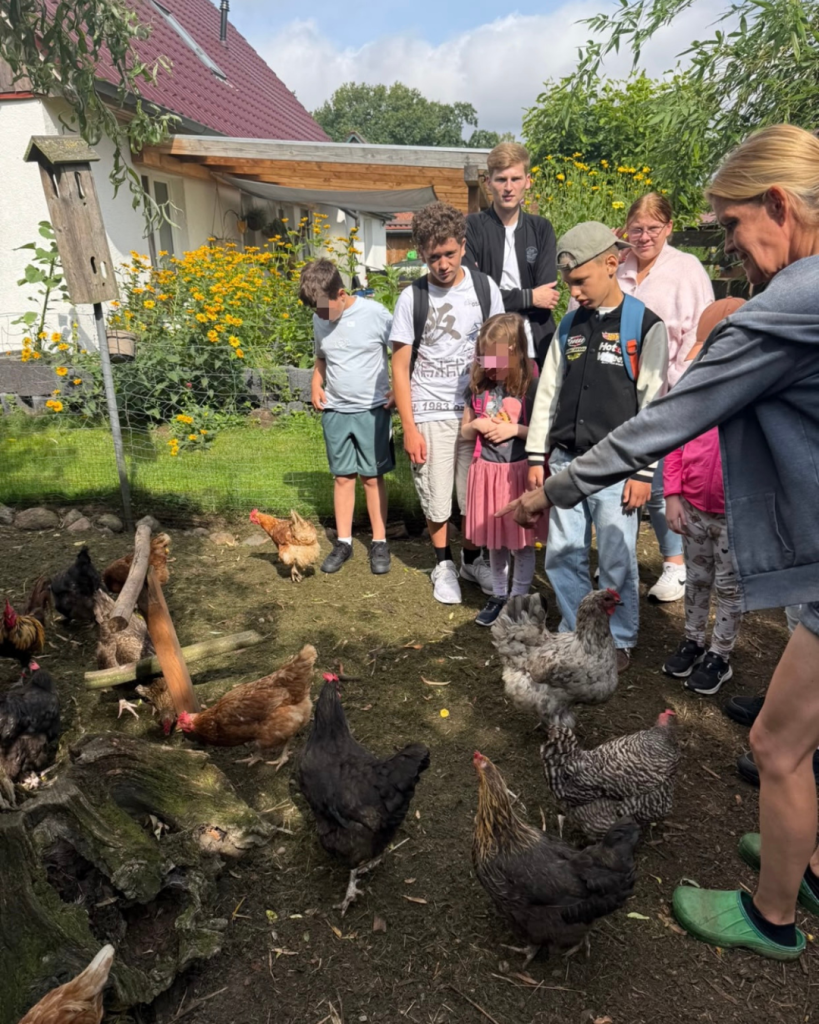 Gruppe von Menschen, darunter Kinder und Erwachsene, die sich im Freien versammelt haben, um Hühner zu beobachten.