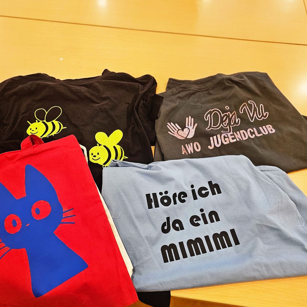Vier T-Shirts und ein Beutel mit verschiedenen Designs auf einem Holztisch ausgelegt.