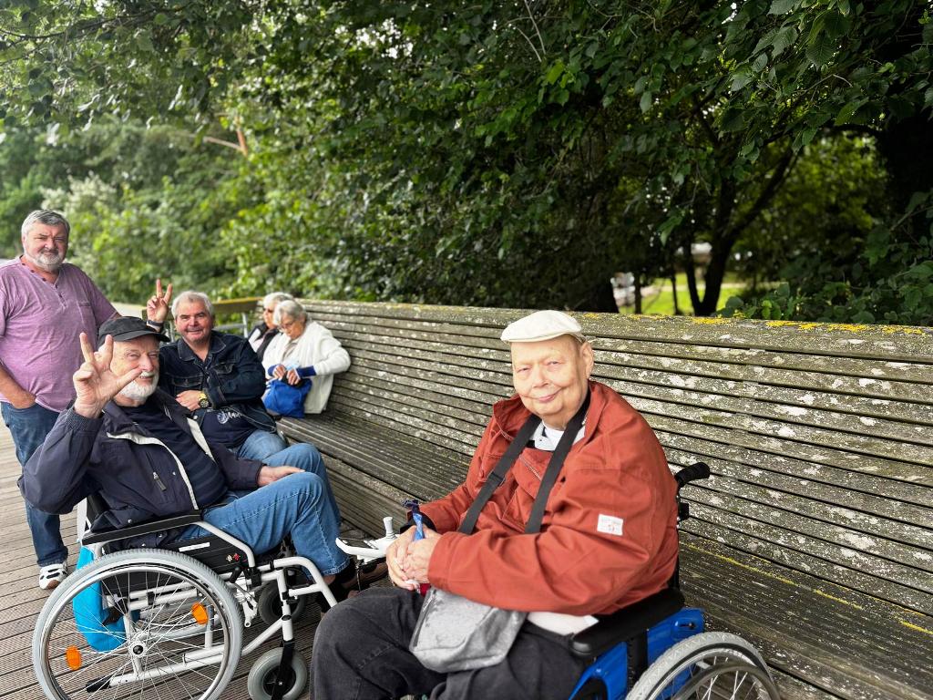 Gruppe von älteren Menschen auf einer Holzbank im Freien, zwei in Rollstühlen