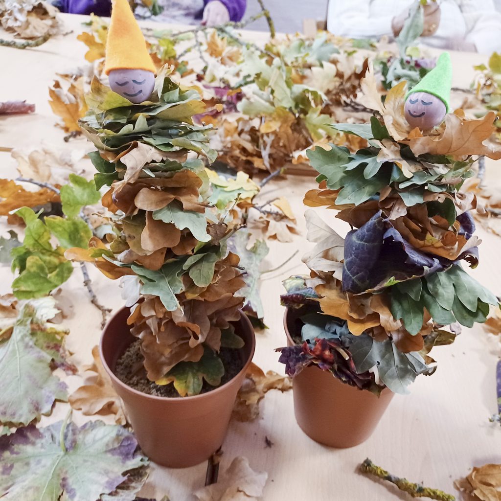 Zwei Herbstdekorationen aus aufeinandergestapelten bunten Blättern in Tontöpfen mit kleinen Stoffmützen als Gesichter.