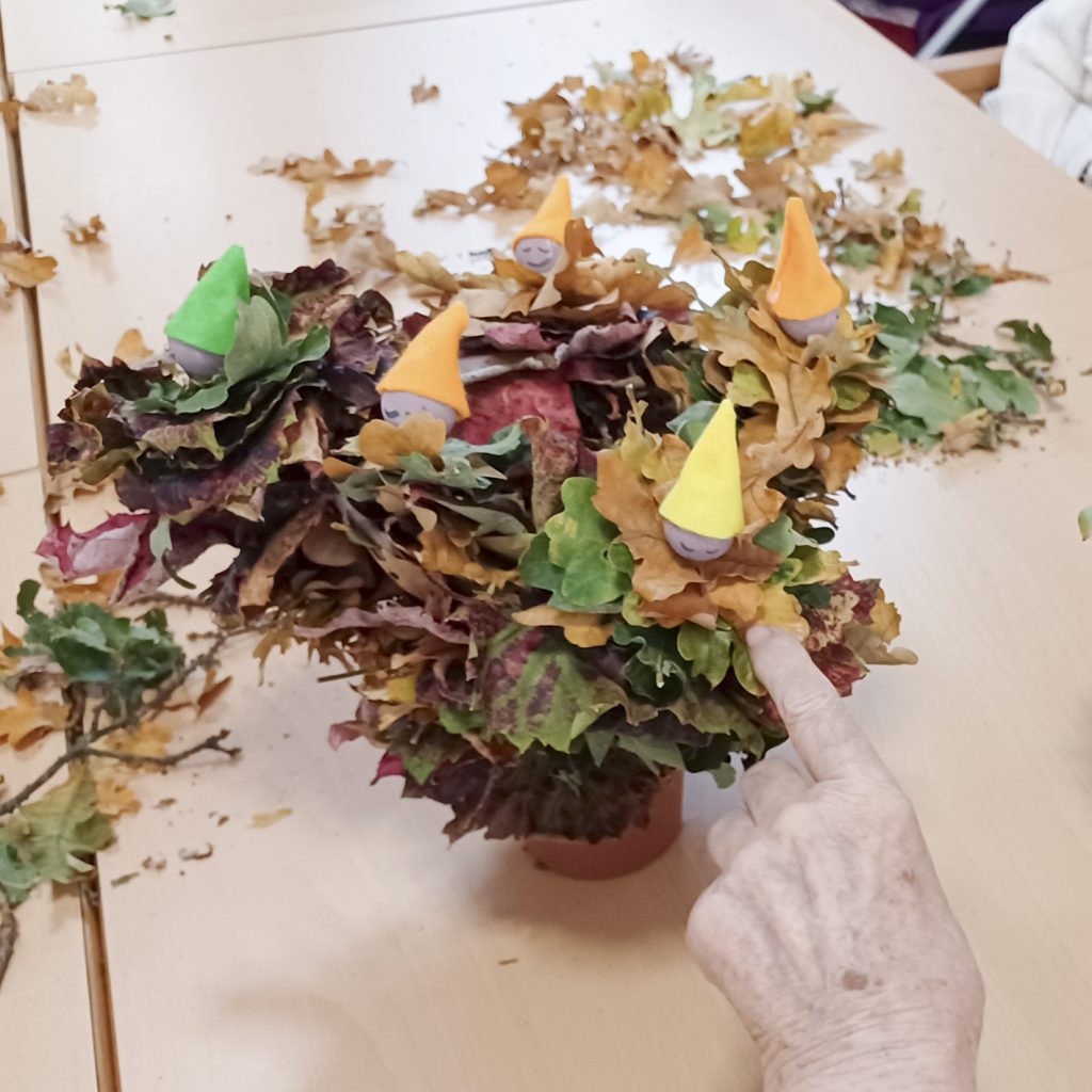 Mehrere bunte Papiermännchen in einem Arrangement aus Herbstblättern auf einem Tisch.