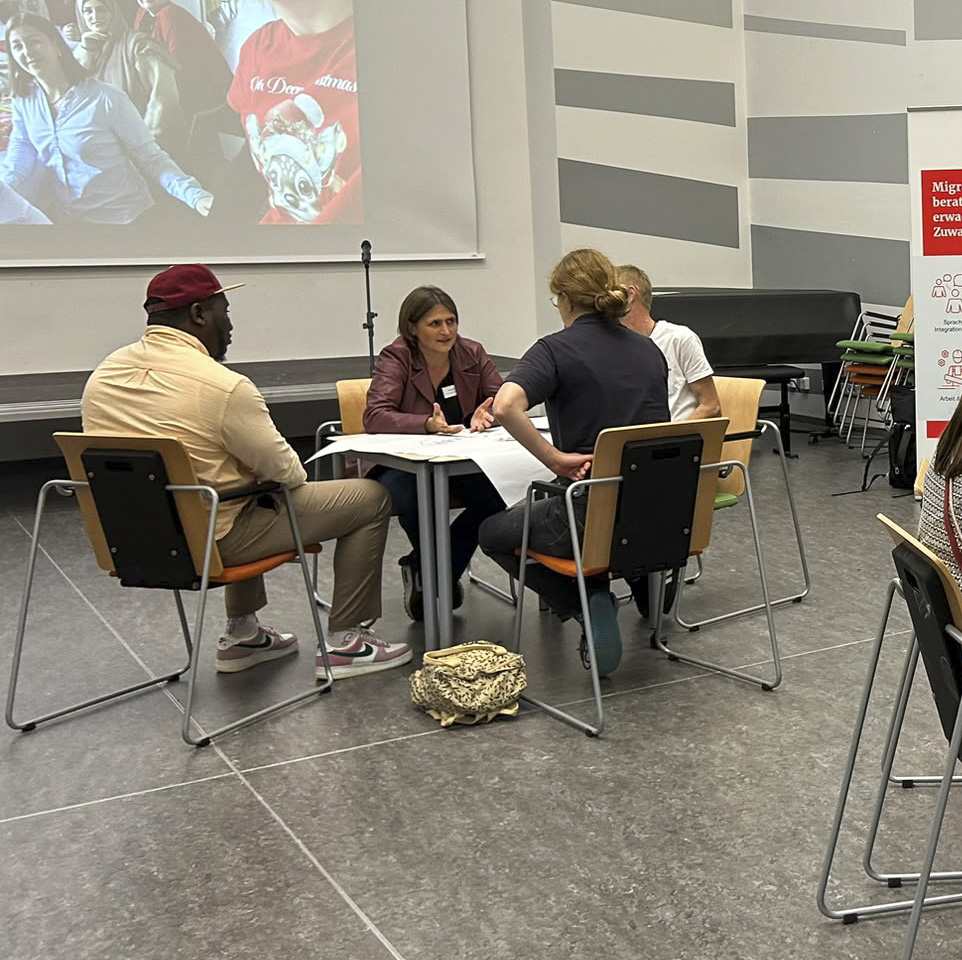 Gruppe von vier Personen, die an einem Tisch in einem Raum sitzt und diskutiert, im Hintergrund eine Projektionswand.