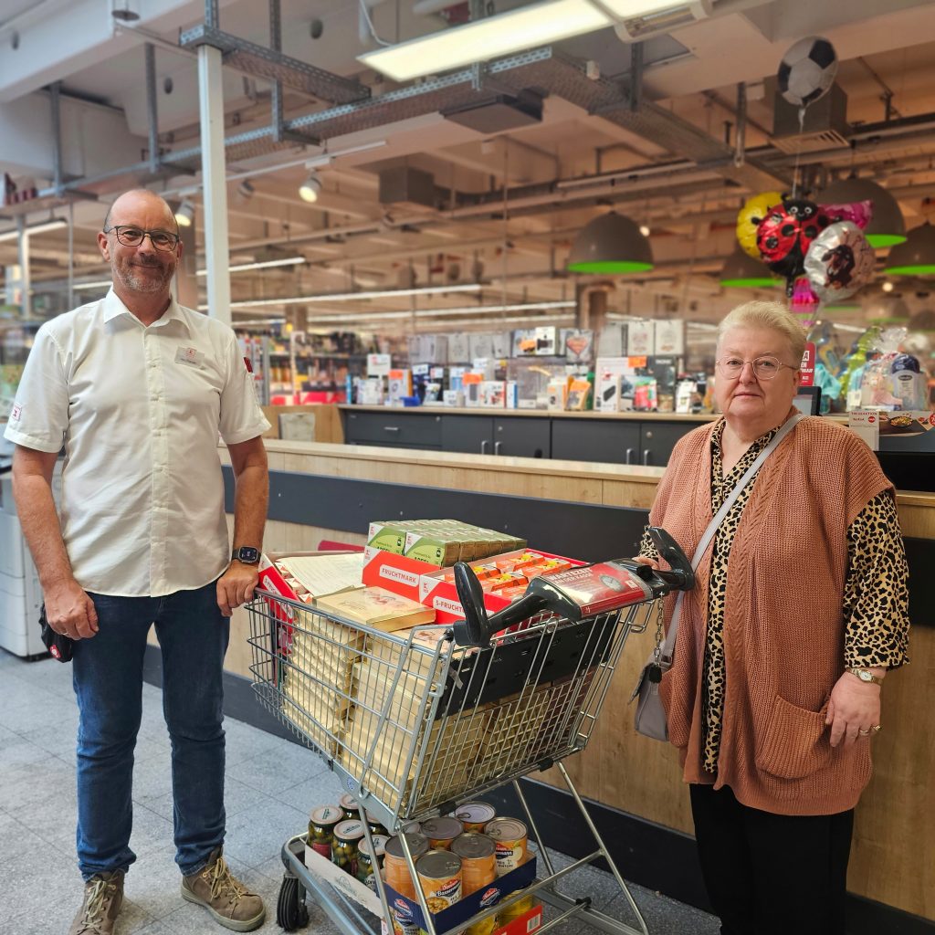 Zwei Menschen stehen neben einem gefüllten Einkaufswagen in einem Laden.