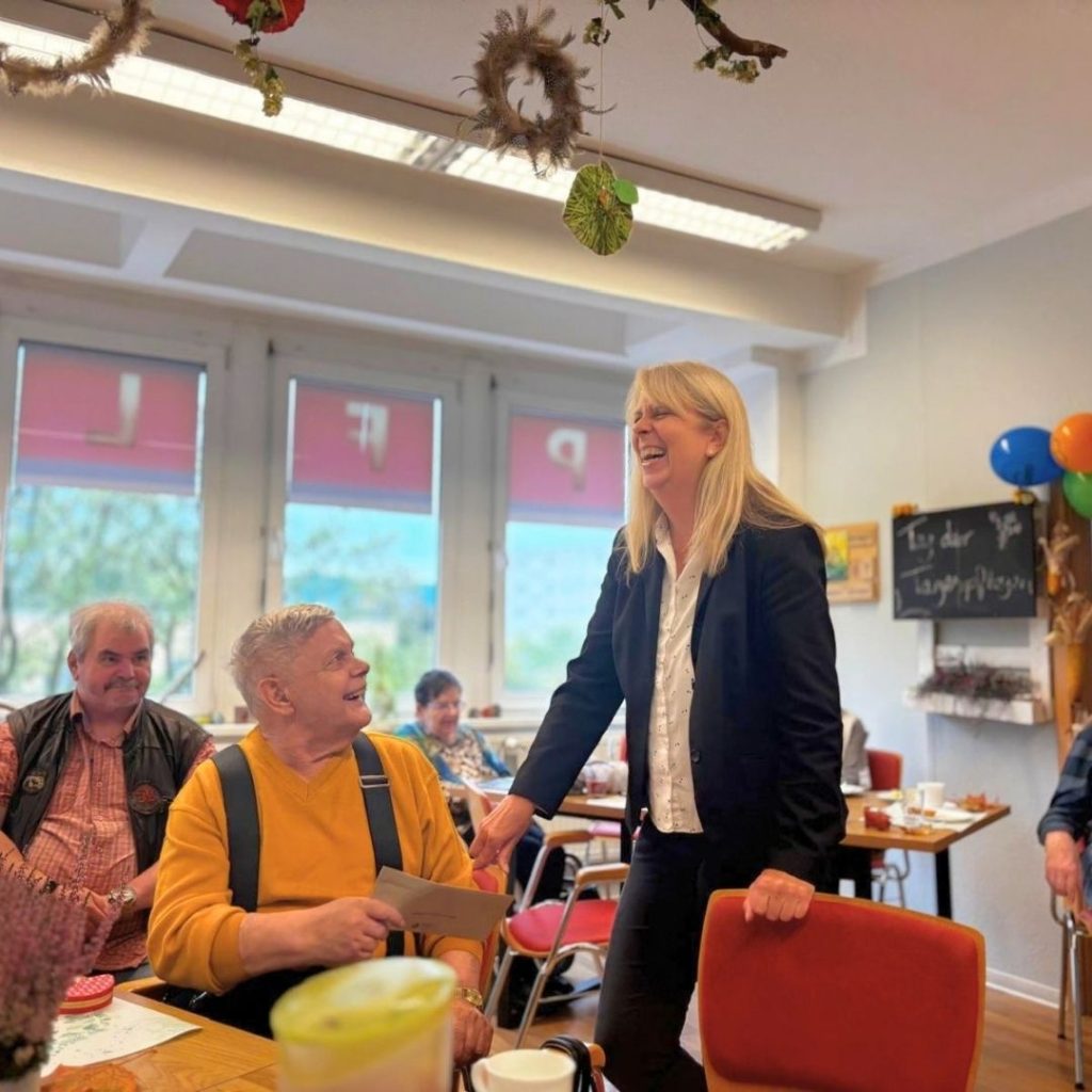Eine Frau im Anzug steht l&auml;chelnd in einem Raum voller Menschen, die an Tischen sitzen. Ein &auml;lterer Mann in einem gelben Pullover sitzt nahe bei ihr und l&auml;chelt. Der Raum ist hell mit gro&szlig;en Fenstern und Dekorationen an der Decke.