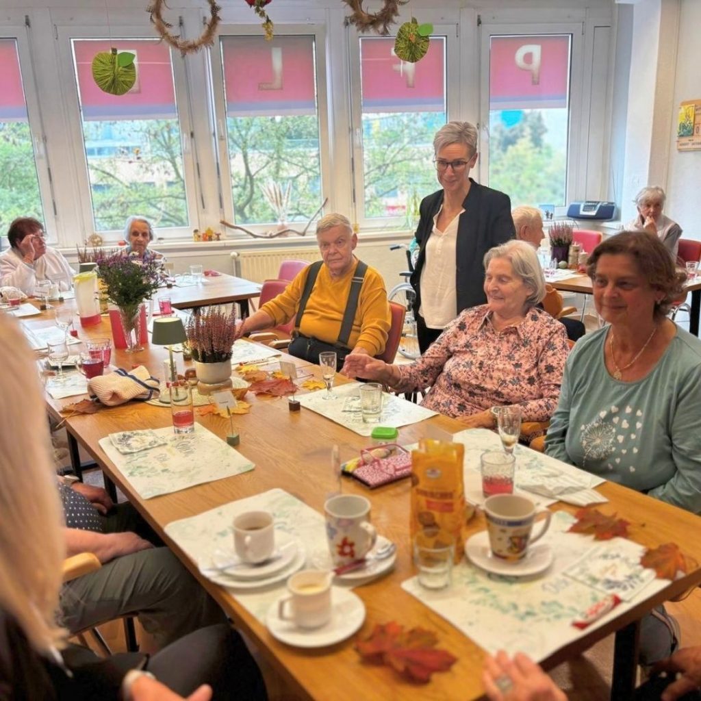 Gruppe von Personen bei einem geselligen Beisammensein in einem Raum mit gro&szlig;en Fenstern.